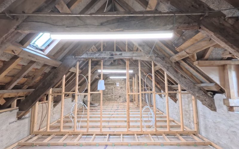 The top floor of Island House today: dark historic roof trusses, purlins and rafters standing open above a long room, with new stud partitions framed up beneath them.